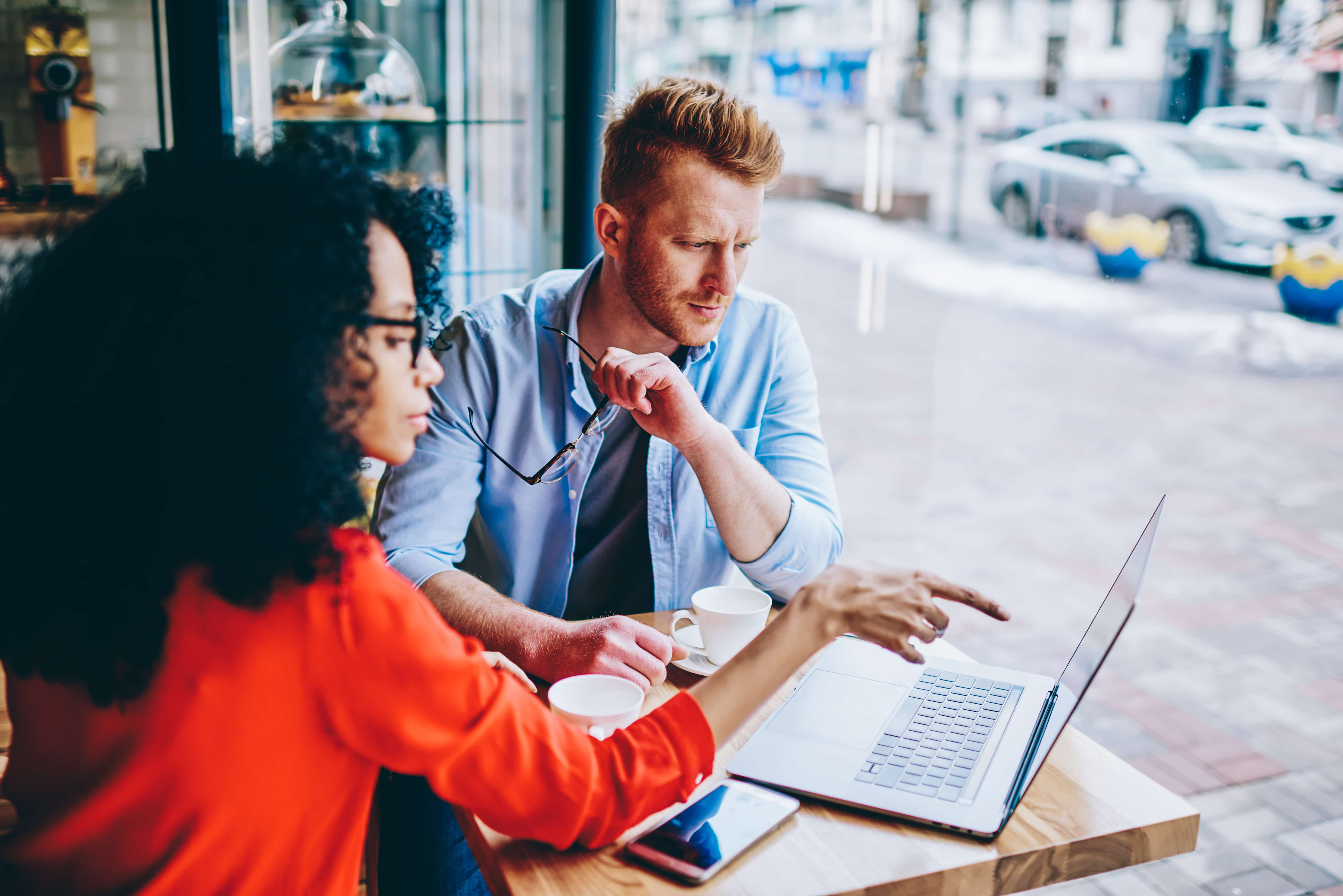 Serious african american female manager pointing with finger on laptop computer showing information with data of financial profit pensive caucasian colleague during business meeting in cafeteria